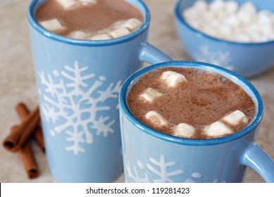 Snowflake mugs filled with hot chocolate and marshmallows on tile counter with cinnamon sticks along side.  Matching bowl with marshmallows in soft focus in background.  Closeup with shallow dof. - Powered by Shutterstock