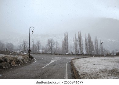 Snowflakes gently fall over Vall de Boí, creating a tranquil winter landscape. Leafless trees line the road while a soft white coating covers the ground and surroundings. - Powered by Shutterstock