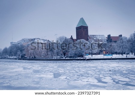 Snow-laden trees and icy serenity define winter in Berlin. A stone church, bathed in warm brown hues, stands as a historic centerpiece amidst the pristine snowy landscape.
