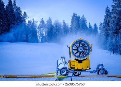 A snowmaking machine is actively producing artificial snow at a ski resort. The surrounding area features evergreen trees and a snowy landscape under a blue sky.