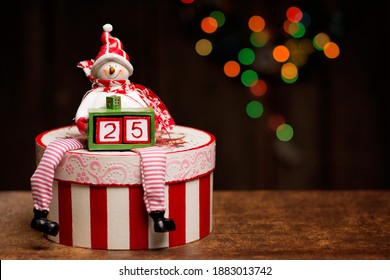 A snowman figure is sitting on a beautifully decorated decoupage box and holding a smaller box with two cubes forming a number twenty-five. A beautiful bokeh made with Christmas lights is in the 