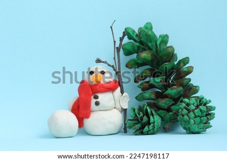 Snowman made of modelling clay with pepper eyes and twig broom. Pinecones painted green . Lightblue background. Wintery set-up.