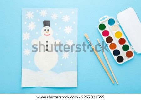 Snowman and snowflakes draw on paper, paint brushes, colorful palette on light blue table background. Pastel color. Closeup.