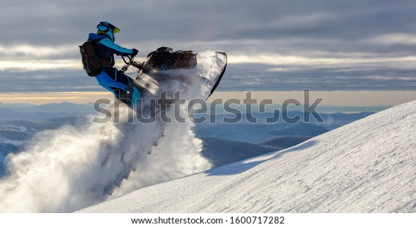 a
snowmobile rider jumps in a mountain valley at dawn. sports snow
bike with snow splashes and snow trail. bright snowmobile and suit
without brands. snowmobilers sports riding. stock
photo