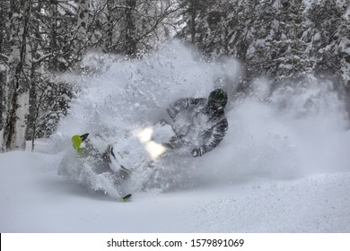 Snowmobile Rider Makes A Turn And Jump During A Snowfall. In The Winter Forest, Leaving Behind A Trail Of Big Splashes Of White Snow From A Snow Bike. Bright Snowmobile And Black Suit. Snowmobilers