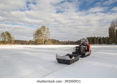 Snowmobile With A Trailer Is On The Lake, Around The Snowy Forest In Sunny Weather