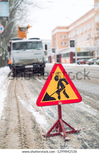 Snow-plow remove snow from the city street.Warning\
road sign.Winter service vehicle snow blower work.Cleaning snowy\
frozen roads