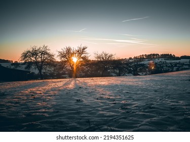 A Snowy Field With Rural Houses In The Background At Sunset