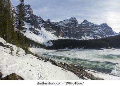 Snowy Hiking Trail Around Frozen Ice Covered Moraine Lake And Scenic Canadian Rocky Mountains Early Springtime Landscape With Alpine Mountain Peaks On Horizon