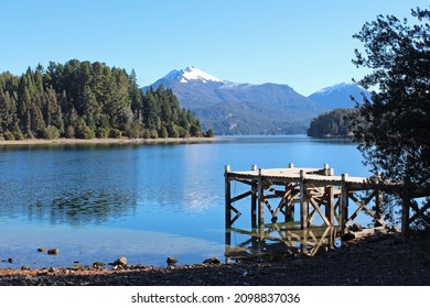 Snowy mountain panorama on Victoria Island, varied vegetation, conifers, old pier, calm Nahuel Huapi lake and clear blue sky. - Powered by Shutterstock