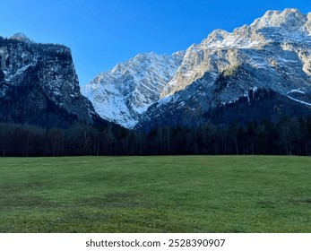 Snowy mountain range with a forested base viewed on a sunny day from Königssee across a green grass field near Saint Bartholomew's Church, Germany. A stunning alpine landscape.