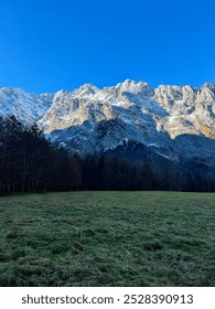 Snowy mountain range with a forested base viewed on a sunny day from Königssee across a green grass field near Saint Bartholomew's Church, Germany. A stunning alpine landscape.