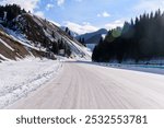 Snowy mountain road winding between snow-covered slopes and pine forests under a clear blue sky.