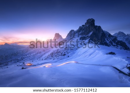 Snowy mountains and blurred car headlights on the winding road at night in winter. Beautiful landscape with snow covered rocks, house, mountain roadway, blue starry sky at sunset in Dolomites, Italy 