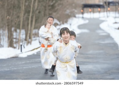 Snowy path lined with bare trees. Family dressed in traditional white robes. Children smile as they run together. Parents carry toddlers with joyful energy. Winter scene evokes warmth and togetherness