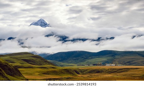 Snowy peak veiled by clouds, green grassland, monastery nestled among rolling hills, dramatic sky. - Powered by Shutterstock