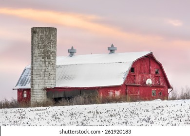 A snowy red barn with silo and basketball hoop is seen with a sunset sky of soft colors.