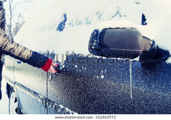 Snowy winter in a city on a sunny day. Car after\
snowfall in the parking lot. Young woman trying to open the icy\
car\
                             \
