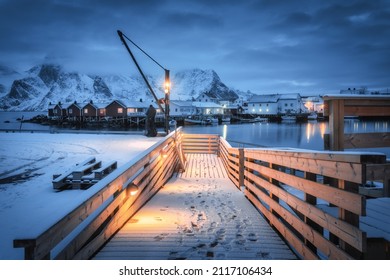 Snowy Wooden Pier On The Sea Coast With Lights, Rorbu And Houses, Boats And Snow Covered Mountains In Clouds At Night. Landscape With Jetty, Buildings, Rock In Fishing Village. Lofoten Islands, Norway