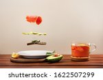 Soaring ingredients for avocado toast. Smoked salmon, avocado slices, fresh basil leaves and dark rye bread floating in air above plate. On table cup of tea, half an avocado. Levitation food concept. 