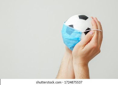 Soccer Ball With A Medical Mask In Hands Of Man On A Gray Background. Cancellation Of Sporting Events. Copy Space.