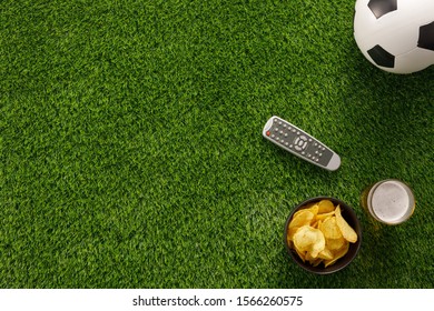 Soccer Ball On A Green Field And Ottoman For A Fan With Snacks And A TV Remote Control. Flat Lay. The Concept Of Football Matches.