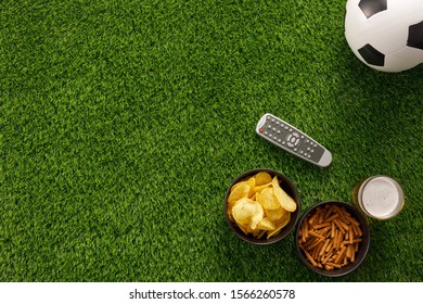Soccer Ball On A Green Field And Ottoman For A Fan With Snacks And A TV Remote Control. Flat Lay. The Concept Of Football Matches.