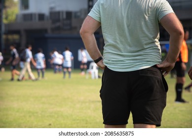 Soccer Dads Standing And Watching Their Sons Playing Football In A School Tournament On A Clear Sky And Sunny Day. Sport, Outdoor Active, Lifestyle, Happy Family And Soccer Mom  Soccer Dad Concept.