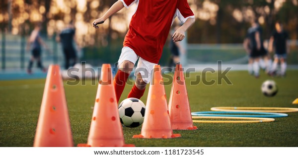 Soccer Drills: The Slalom Drill. Youth soccer\
practice drills. Young football player training on pitch. Soccer\
slalom cone drill. Boy in red soccer jersey shirt running with ball\
between cones