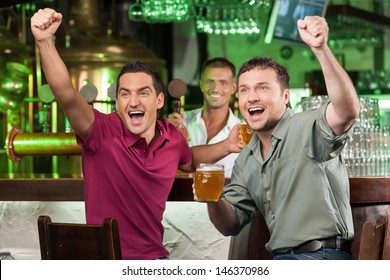 Soccer Fans At The Bar. Two Happy Football Fans Cheering At Bar And Drinking Beer While Bartender Serving Beer At The Background