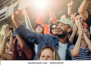 Soccer fans taking a selfie at stadium during a match with fellow supporters around.  Group of supporters watching a match and cheering. - Shutterstock ID 1650156529