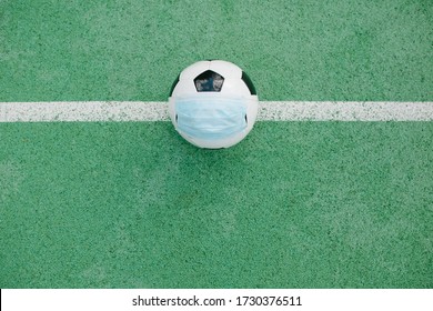 Soccer Football With Surgical Face Mask On Middle Line Of Artifiical Turf Field, Ready For Kick Off, Symbol Image For Sports During The Corona Covid-19 Pandemic In 2020