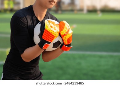 A soccer goalkeeper in a black jersey is holding a soccer ball with both hands, wearing vibrant orange and yellow goalkeeper gloves.  - Powered by Shutterstock