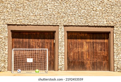 Soccer Net And Two Balls Set Up In Driveway In Front Of Wooden Doors Of  Two Car Garage Of Rock House