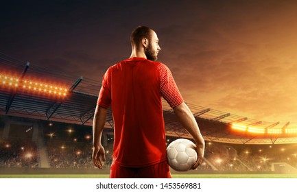 Soccer Player With A Ball Stands In Front Of Cheering Fans On The Stadium. View From Back. Floodlit Soccer Stadium With Dramatic Sky. 