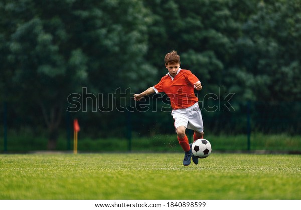 Soccer shooting. Boy kicking soccer ball on grass\
field. Young football player in action running jumping, and\
shooting the ball. Junior level sports competition. Footballer in\
red jersey shirt