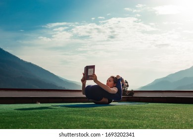 Sochi, Russia 03.26.2020 Attractive young woman performing yoga on the green lawn and reading a book in a funny position with legs over her head with great mountain view background