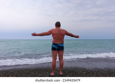 Sochi, Russia - January 24, 2022: A Man Stands In Blue Swimming Trunks On A Pebble Beach And Dries After Swimming In The Cold Winter Sea. Cold Water, Cloudy Sky. Selective Focus.