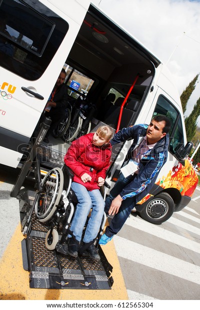 SOCHI, RUSSIA - MAR , 2014: Olympic\
Park. The young man helps the disabled in wheelchairs to enter the\
electric car is equipped for people with\
disabilities.