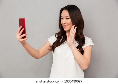 Sociable Beautiful Woman With Asian Appearance Taking Selfie Or Speaking On Video Call Using Cell Phone Isolated Over Gray Background