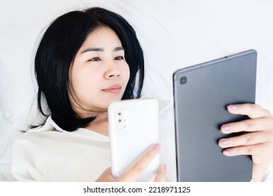 Social Addiction Concept With Asian Woman Holding Smart Phone And Tablet Laying Down In Bed Looking At Screen  