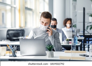 Social Distance And Online Work With Client Remotely. Busy Man In Protective Mask Talking On Smart Phone Sitting At Office In Workplace With Laptop And Glass Board During Coronavirus, Copy Space