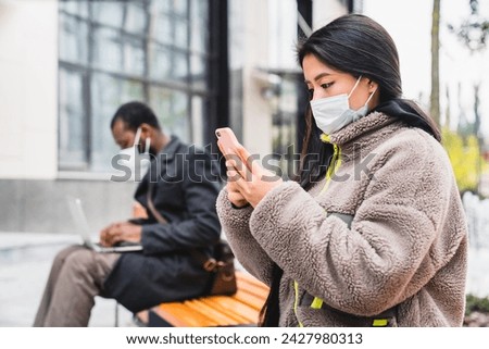 Social distancing concept. Asian girl using phone in medical mask while african man in mask using laptop outdoors. Coronavirus pandemic spread prevention