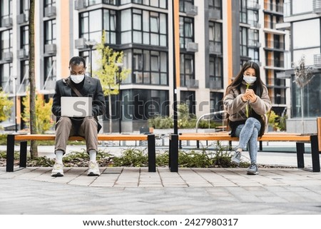 Social distancing concept. Two people in medical masks sitting on benches with laptop and phone outdoors. Coronavirus pandemic spread concept