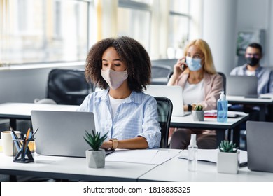 Social Distancing At Coronavirus Outbreak Situation. Pretty African American Lady In Protective Mask Typing On Laptop, Working For Company Support Center In Office Interior With Other Employees