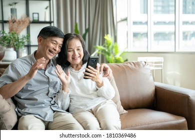 Social distancing or new normal concept.Senior asian couple sitting on sofa in home playing smartphone and video conference with grandchild.They are smiling to spend their time together with happiness