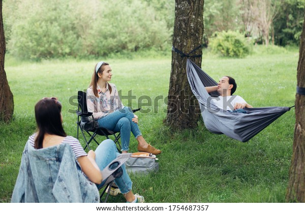 Social distancing. Small group of people\
enjoying conversation at picnic with social distance in summer\
park. Friends chilling in hammock and chairs among trees. New\
normal, safety\
gatherings