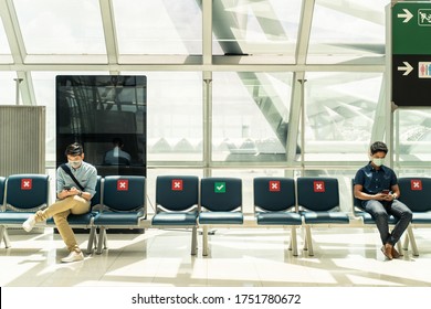 Social Distancing, Two Men Wearing Face Mask Sitting Keeping Distance Away From Each Other To Prevent Covid19 Infection During Pandemic. Empty Chair Seat Red Cross Shows Avoidance In Airport Terminal.