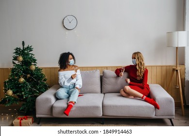 Social distancing while celebrating Christmas and New Years 2021 concept. Two girls in protective masks with glasses of champagne in their hands are sitting on the sofa far from each other.