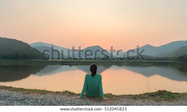 夕暮れの間 女性が湖のそばでのんびり座っている の写真素材 今すぐ編集